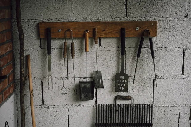 Cooking tools and tongs hanging from a wooden board against a masonry wall